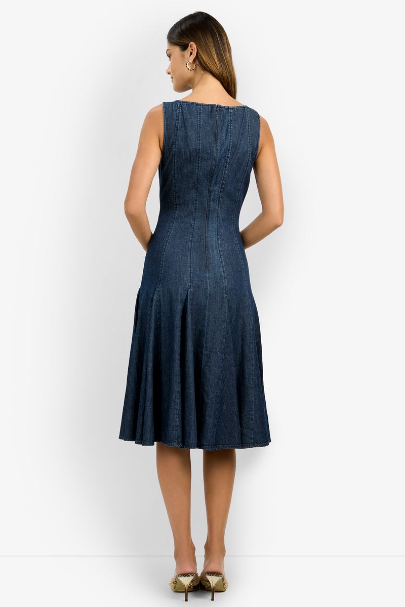 A woman with light brown hair faces away, modeling the sleeveless, knee-length CASIMA DRESS in dark denim with seam details and a flared skirt. She wears ankle-strap pointed-toe shoes against a plain white background.