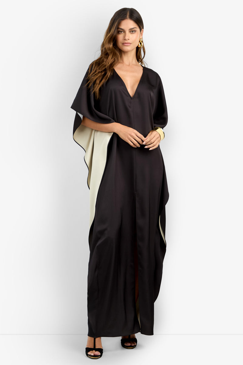 She wears the RIVA DRESS, a long black gown with cream side accents, deep V-neckline, and wide sleeves. Styled with black heels and gold hoop earrings, she poses against a plain white background.