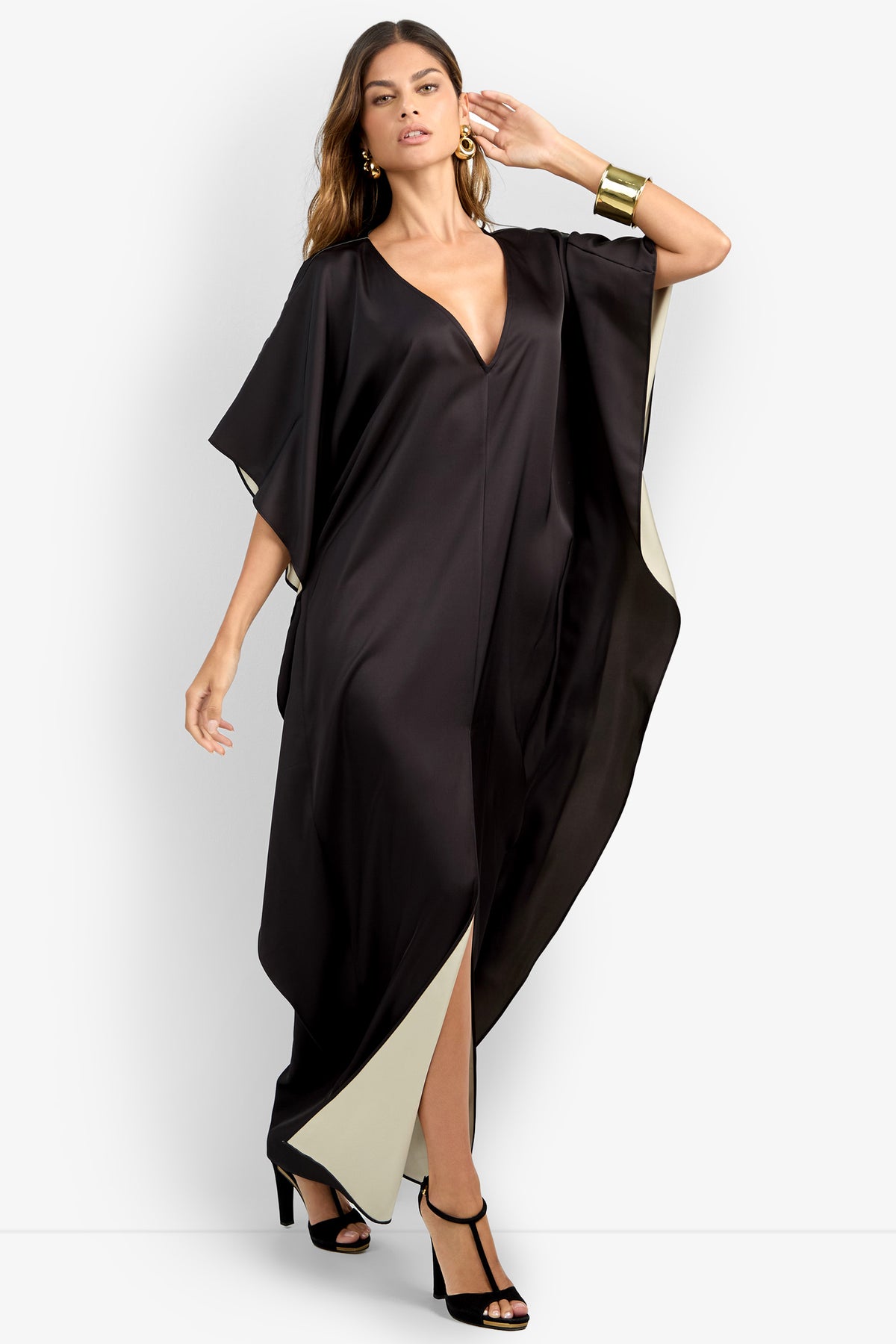A woman models the RIVA DRESS, a black, flowing V-neck kaftan with light lining, paired with black high-heeled sandals, gold earrings, and a wide gold cuff bracelet against a plain white background.