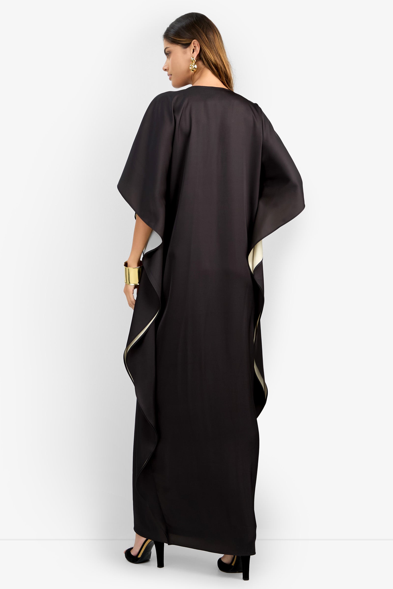 A woman faces away in the RIVA DRESS, a long, loose black dress with wide sleeves and cream trim. She wears black heels, gold cuff bracelet, earrings, and has straight brown hair. The background is plain white.