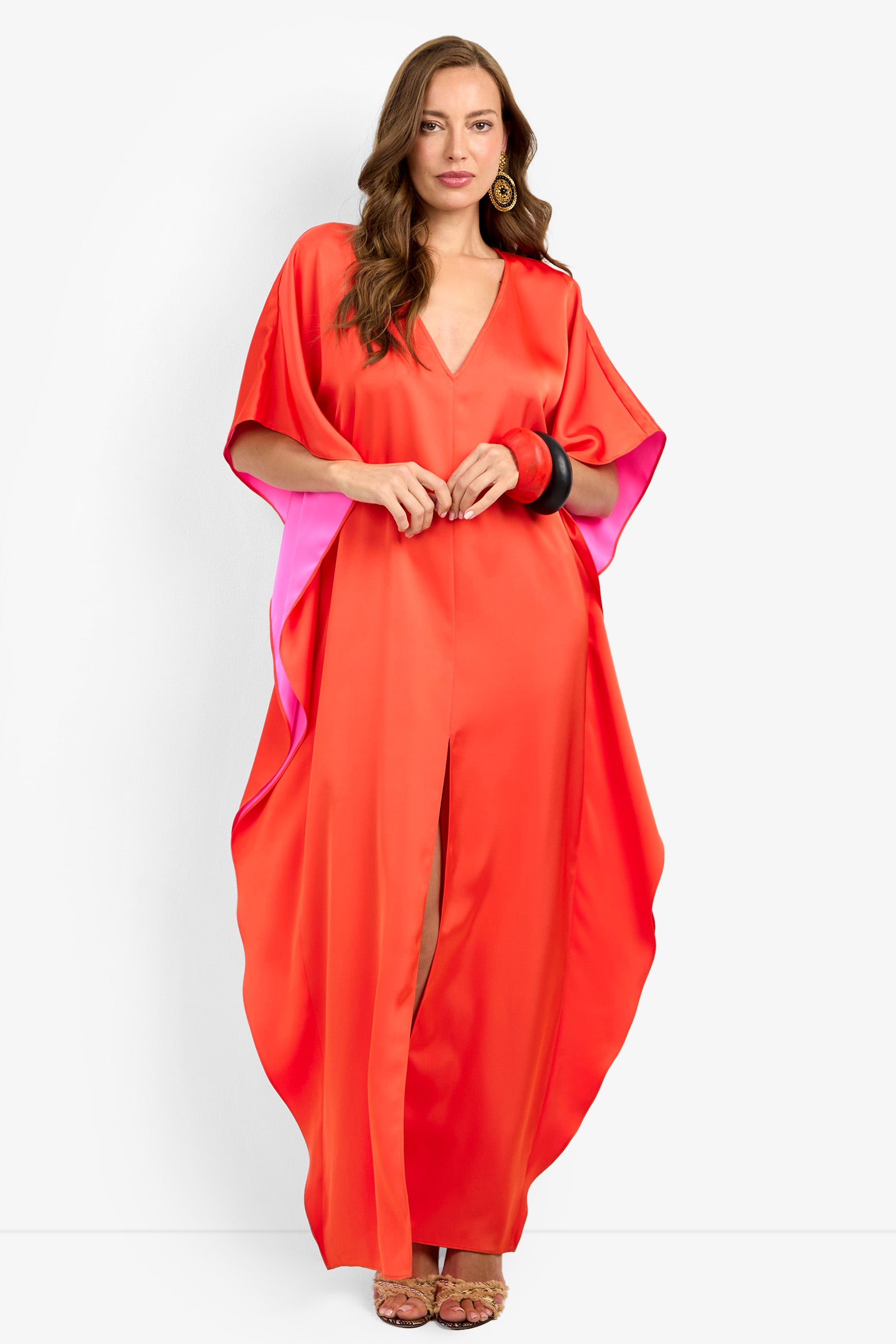 A woman stands facing forward in the loose, bright red-orange RIVA DRESS with pink lining. She has long brown hair, wears gold statement earrings, bold bracelets, and strappy sandals.