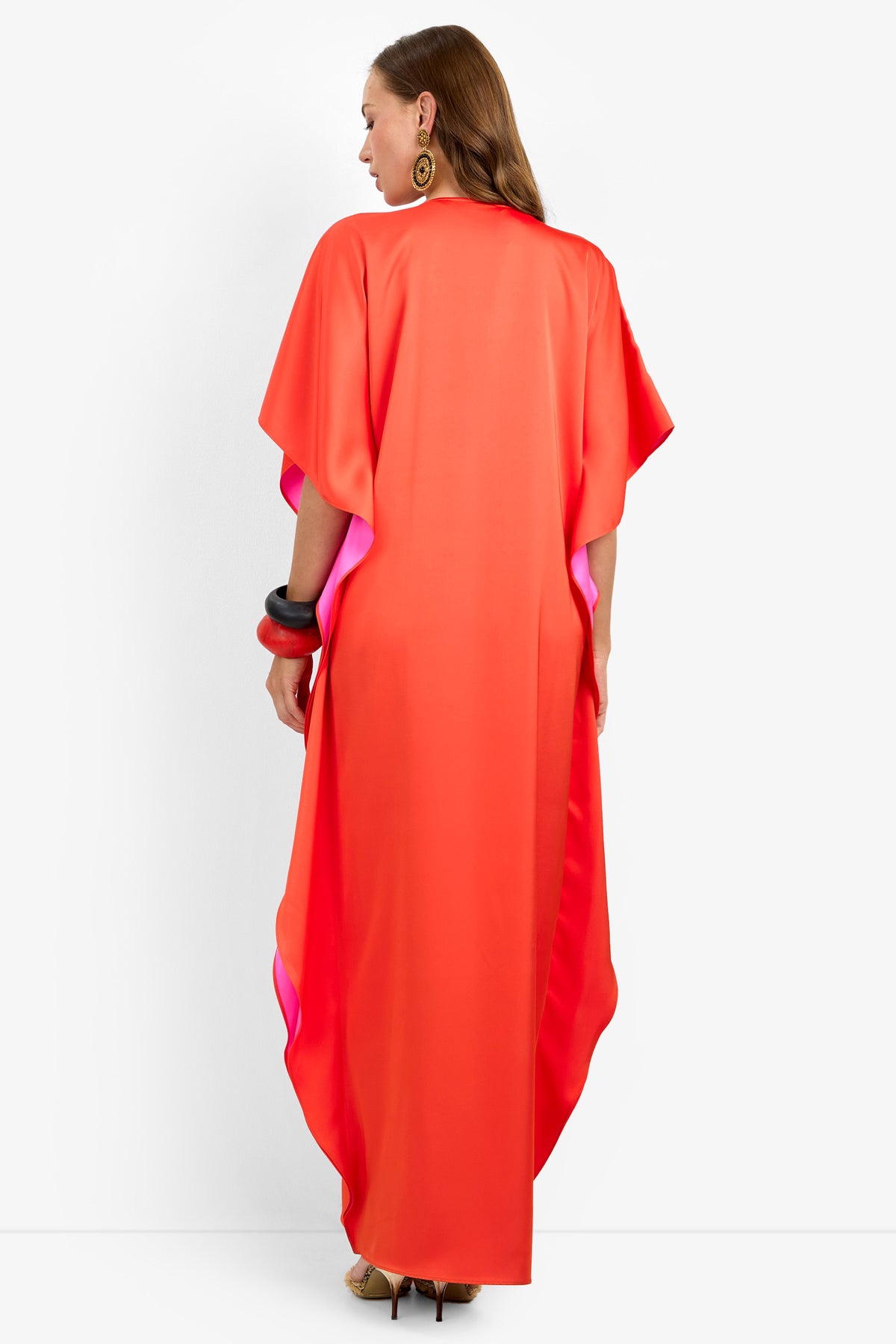 A woman stands facing away, wearing the RIVA DRESS—a long, loose-fitting, bright red and pink kaftan with short sleeves. She accessorizes with large gold earrings, chunky bangles, and beige high heels.