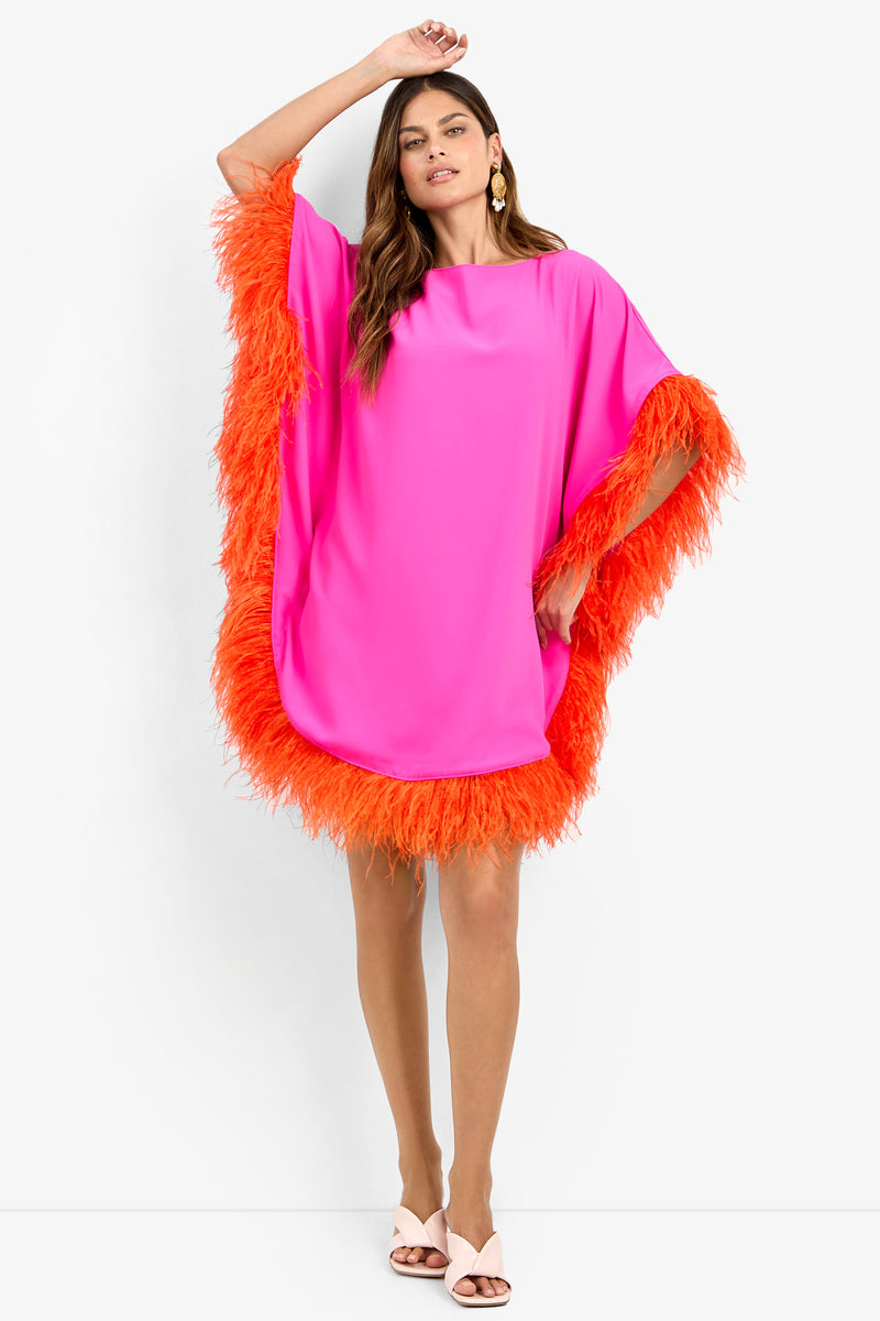 A woman models the MINI NEENA, a loose, hot pink dress with bright orange feather trim on the sleeves and hem. She pairs it with white heeled sandals and gold earrings, her hair in loose waves and one arm raised.