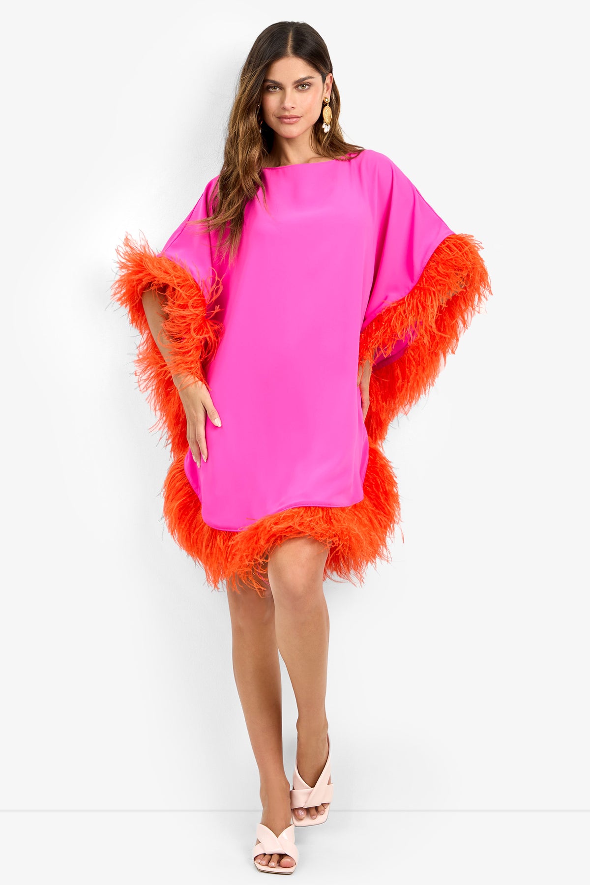 Woman in the MINI NEENA, a loose-fitting bright pink dress with orange feather trim on sleeves and hem, poses against a white background wearing light pink heels and statement earrings.