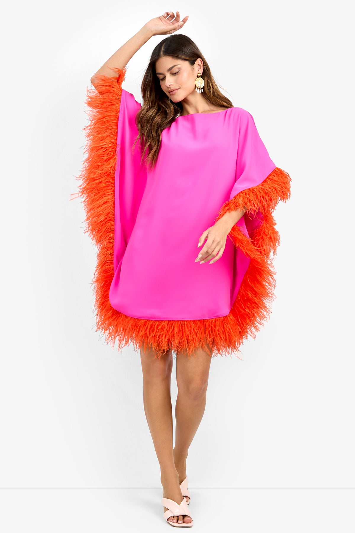 A woman wears the MINI NEENA, a loose-fitting bright pink dress with orange feather trim on the sleeves and hem, styled with large earrings and white open-toe heels, standing against a plain white background.