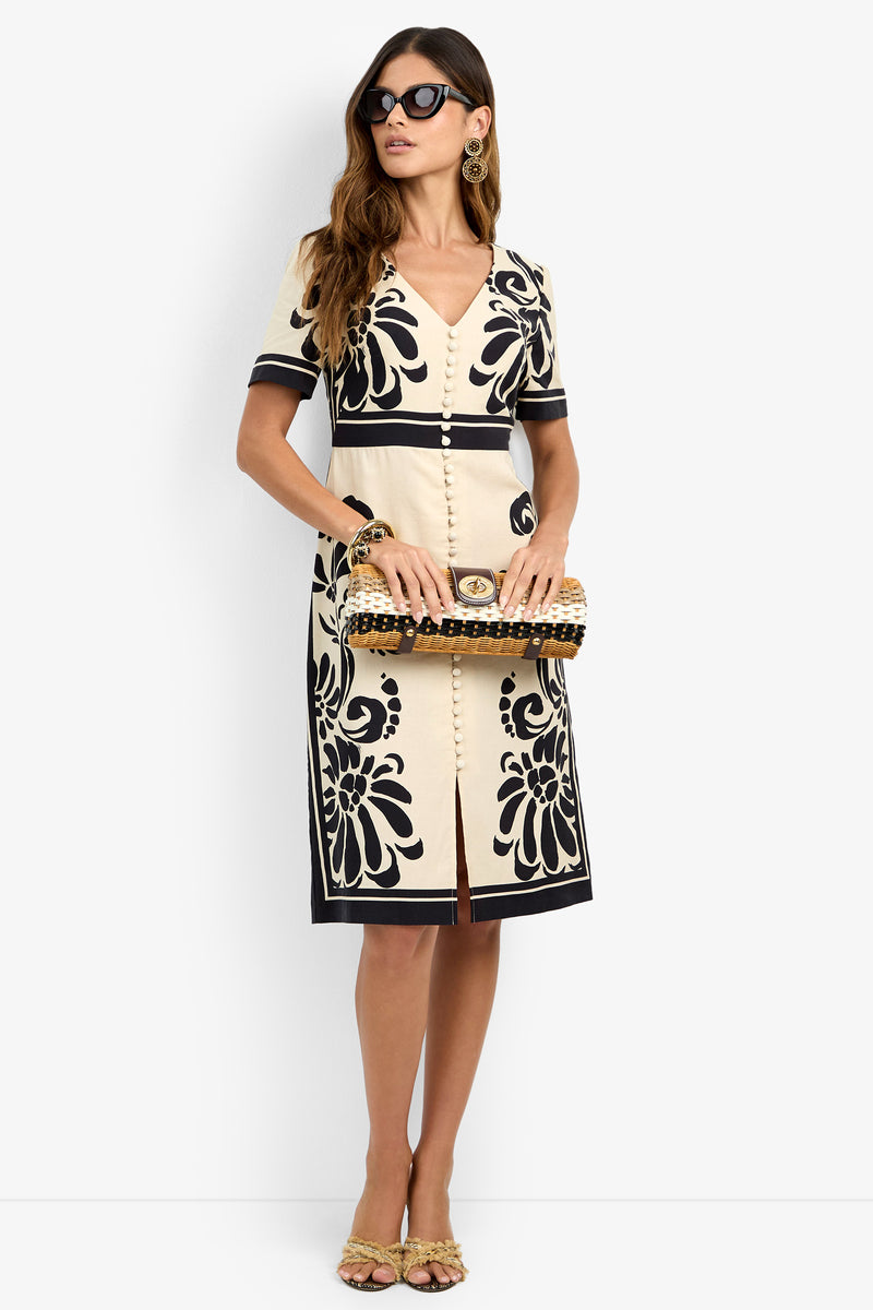 A woman wearing the JENNAH DRESS—cream with bold black floral patterns—paired with sunglasses, a beaded clutch, and heeled sandals, stands against a plain white background.