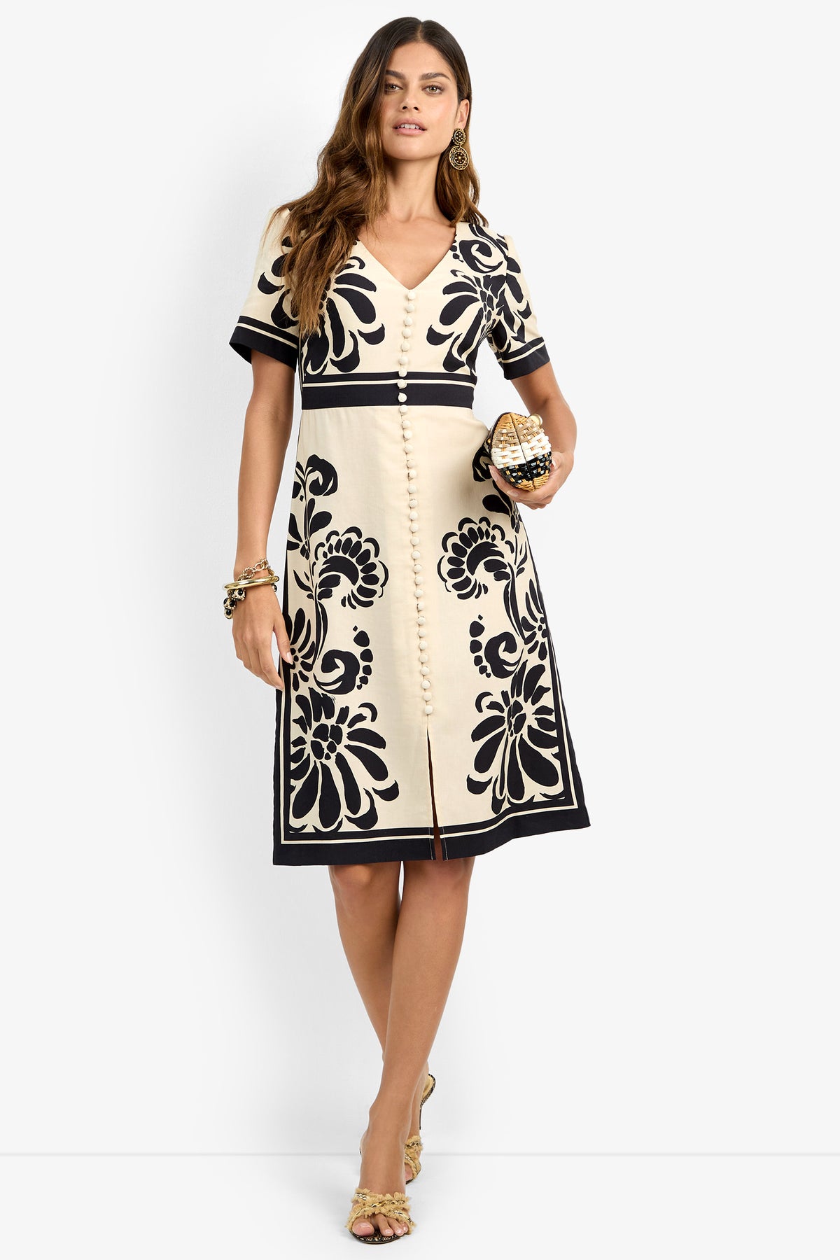 A woman models the JENNAH DRESS, featuring cream fabric with black floral patterns. She accessorizes with gold earrings, a bracelet, and clutch, wearing heeled sandals while standing against a plain white background.