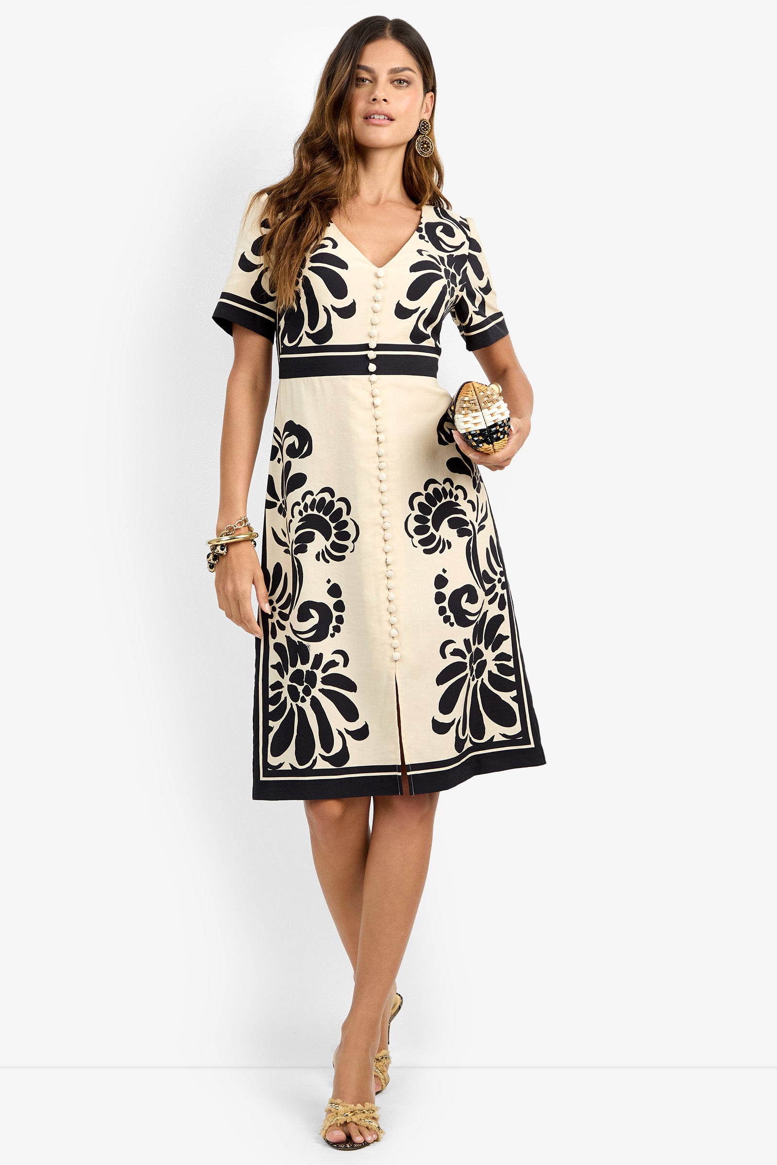A woman models the JENNAH DRESS, featuring cream fabric with black floral patterns. She accessorizes with gold earrings, a bracelet, and clutch, wearing heeled sandals while standing against a plain white background.