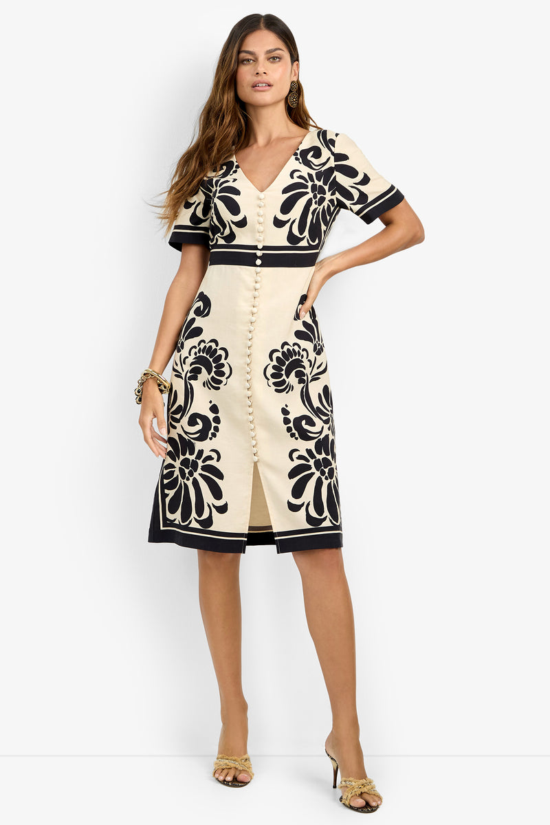 A woman wears the JENNAH DRESS, a cream short-sleeve dress with bold black floral patterns, front buttons, and a small slit at the hem. She accessorizes with gold earrings, a bracelet, and yellow heeled sandals.