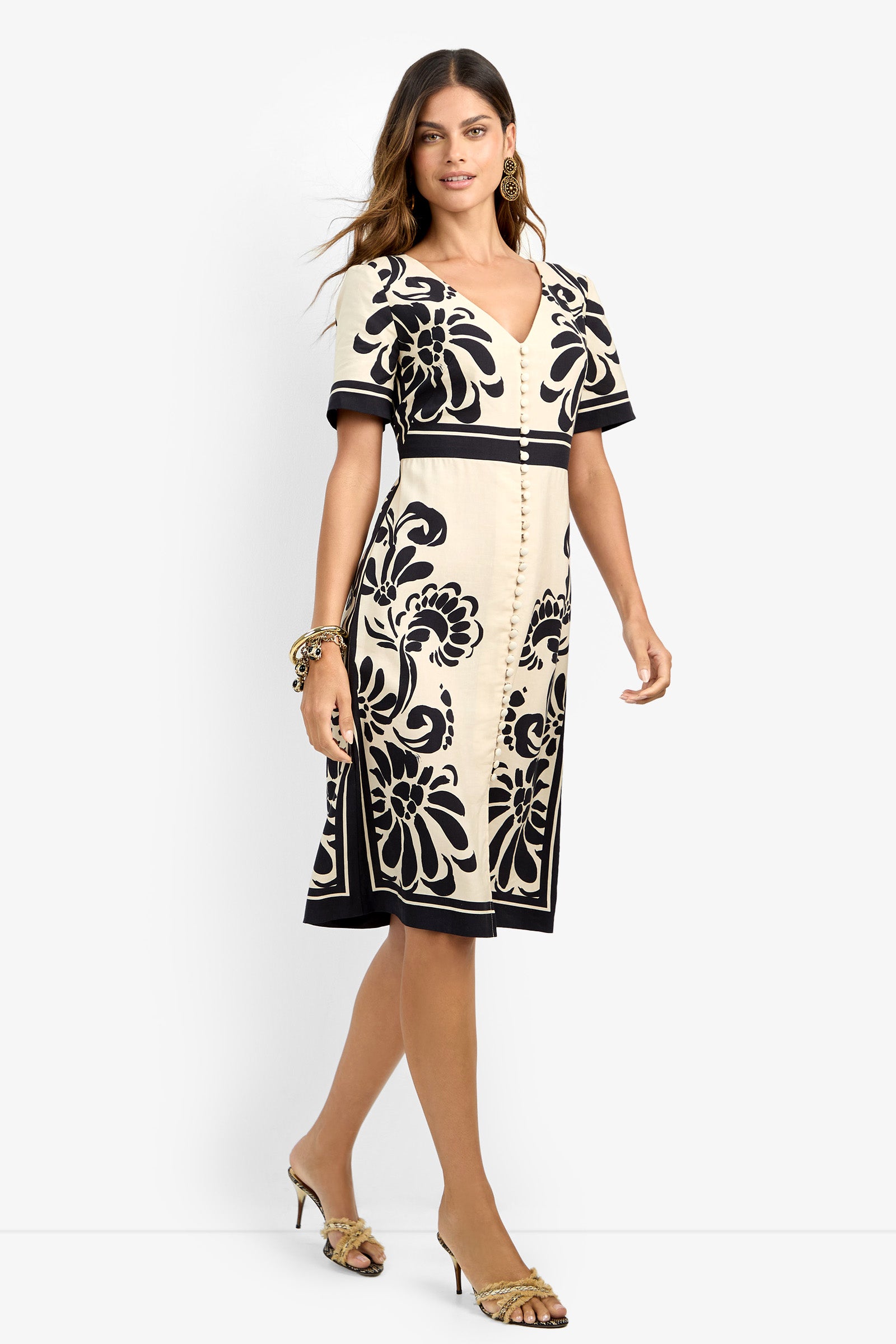 A woman wears the JENNAH DRESS, a cream knee-length dress with bold black floral patterns, short sleeves, and a V-neckline. She completes the look with heeled sandals plus earrings and bracelets.