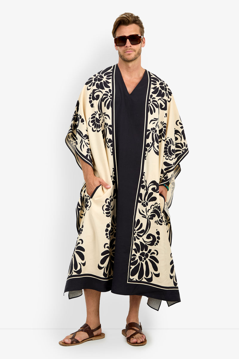 A man in sunglasses and sandals, hands in pockets, wears the SAN MARTINO CAFTAN—a long, dark robe paired with a light floral kimono-style overcoat.