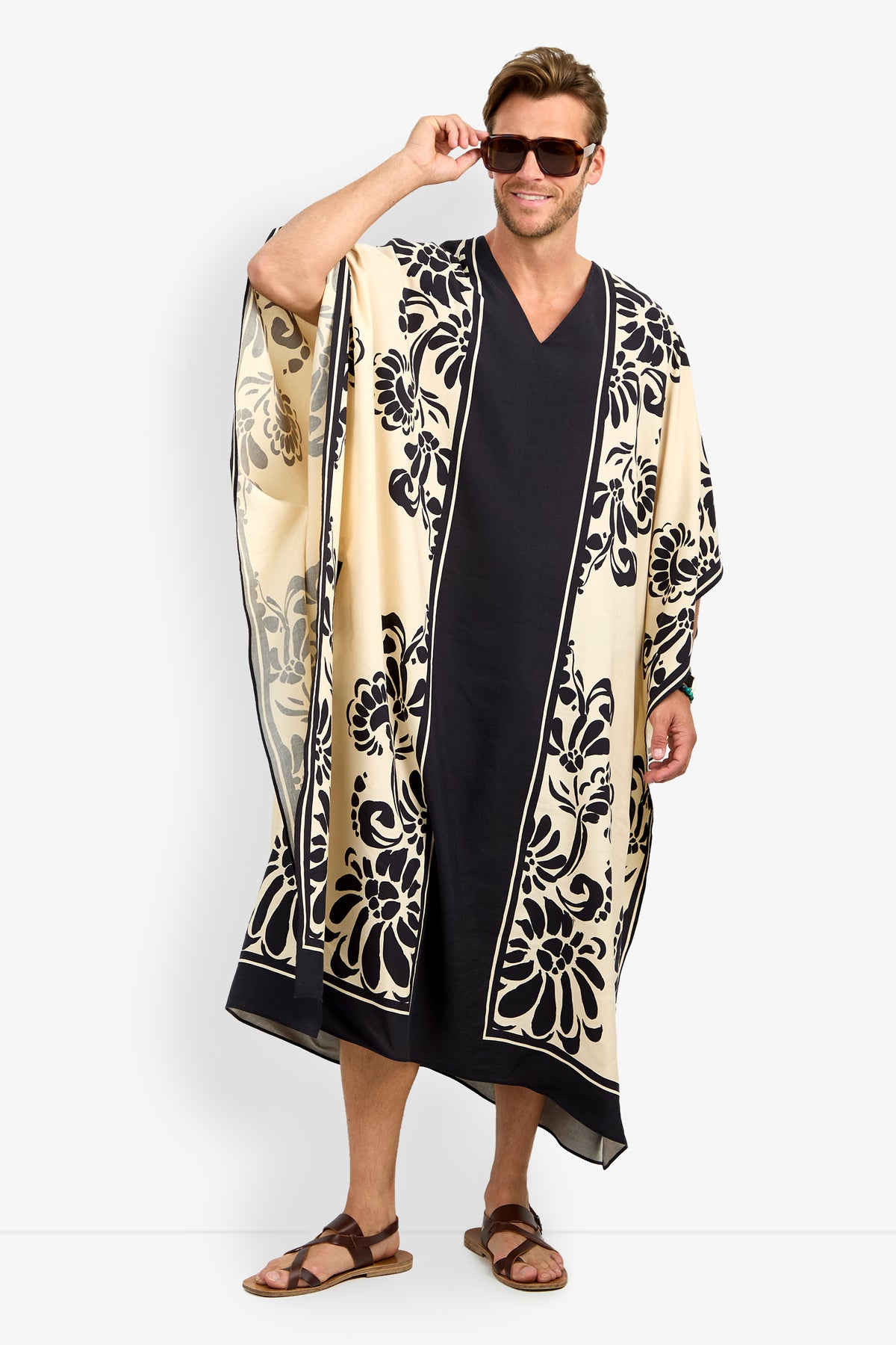 A man in sunglasses and sandals smiles while modeling the SAN MARTINO CAFTAN—a long, loose cream and black garment with floral patterns along the edges and center—standing against a plain white background.