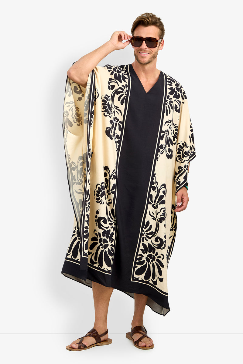 A man in sunglasses and sandals smiles while modeling the SAN MARTINO CAFTAN—a long, loose cream and black garment with floral patterns along the edges and center—standing against a plain white background.