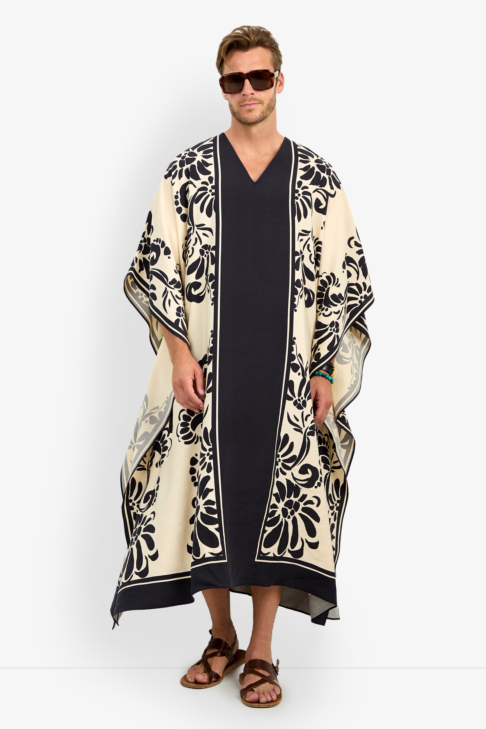A man stands straight in sunglasses and sandals, wearing the SAN MARTINO CAFTAN—a long, loose-fitting cream and navy garment with bold floral patterns on the borders and sleeves.