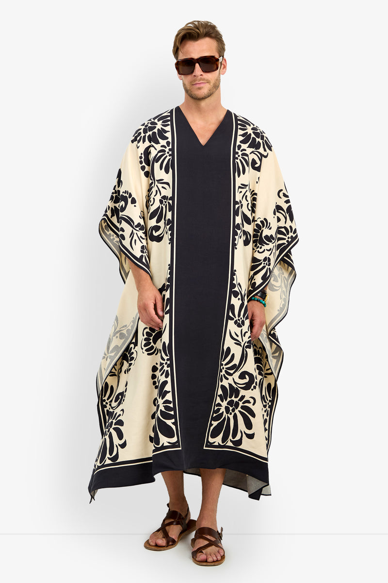 A man stands straight in sunglasses and sandals, wearing the SAN MARTINO CAFTAN—a long, loose-fitting cream and navy garment with bold floral patterns on the borders and sleeves.