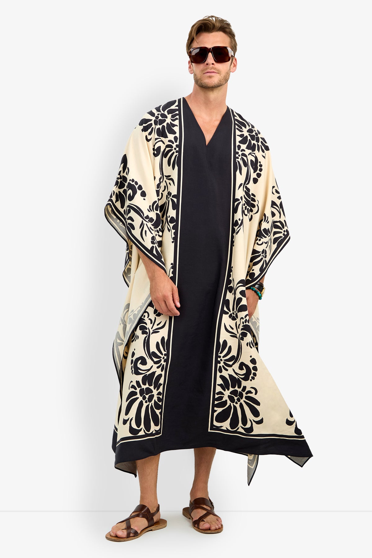 A man faces forward wearing the SAN MARTINO CAFTAN—a long black garment with wide, cream-colored sleeves, ornate black floral borders—paired with sunglasses and sandals.