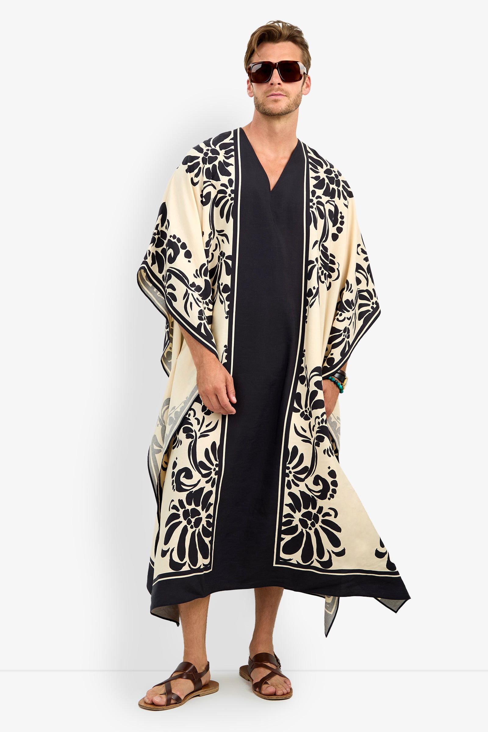 A man faces forward wearing the SAN MARTINO CAFTAN—a long black garment with wide, cream-colored sleeves, ornate black floral borders—paired with sunglasses and sandals.