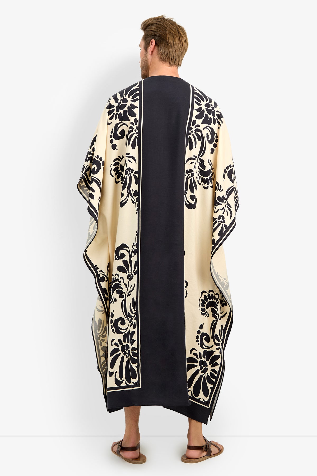 A man with short brown hair and sandals stands against a plain white background, wearing the SAN MARTINO CAFTAN—a long, flowing black and cream garment adorned with bold floral patterns.