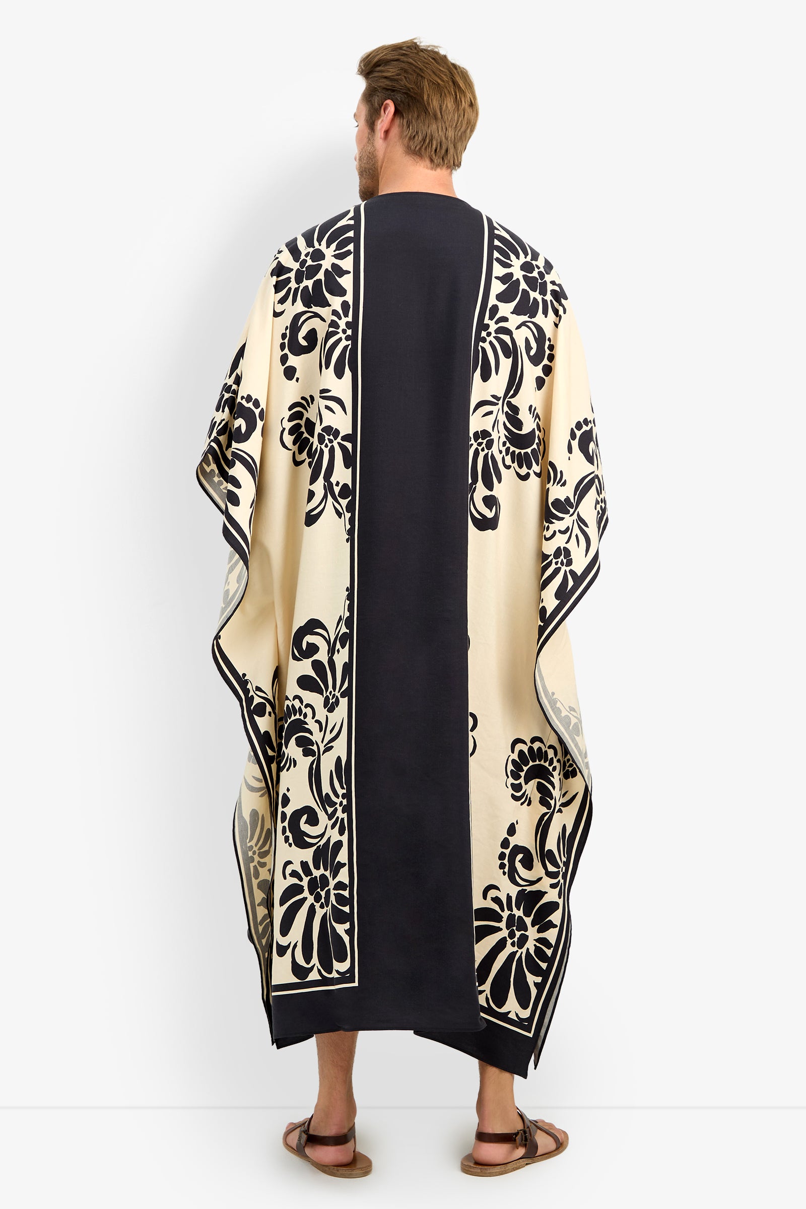 A man with short brown hair and sandals stands against a plain white background, wearing the SAN MARTINO CAFTAN—a long, flowing black and cream garment adorned with bold floral patterns.