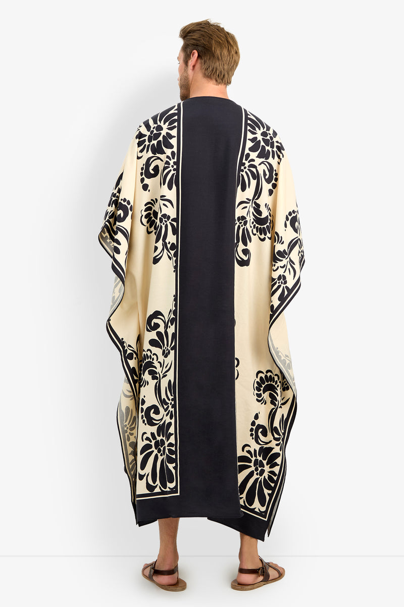 A man with short brown hair and sandals stands against a plain white background, wearing the SAN MARTINO CAFTAN—a long, flowing black and cream garment adorned with bold floral patterns.
