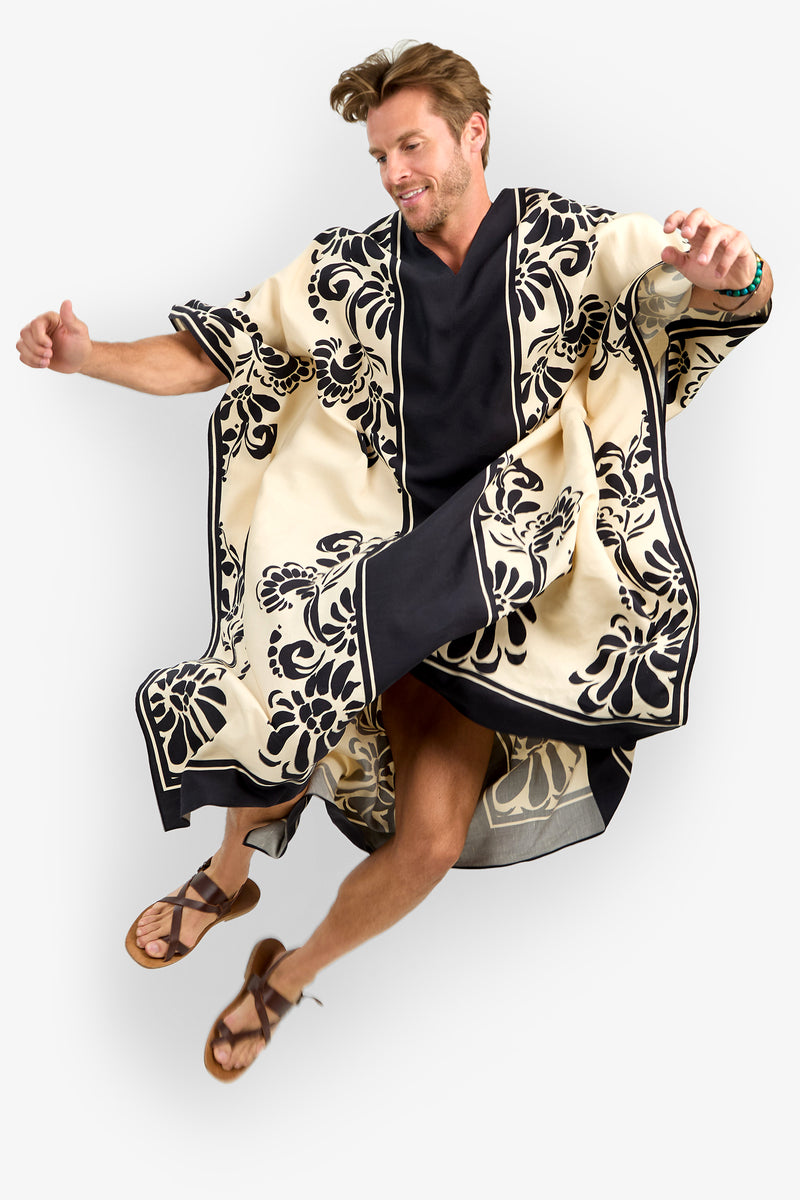 A man wearing the SAN MARTINO CAFTAN, a cream and black floral patterned robe with wide sleeves, smiles while jumping in brown sandals and a black shirt against a white background.