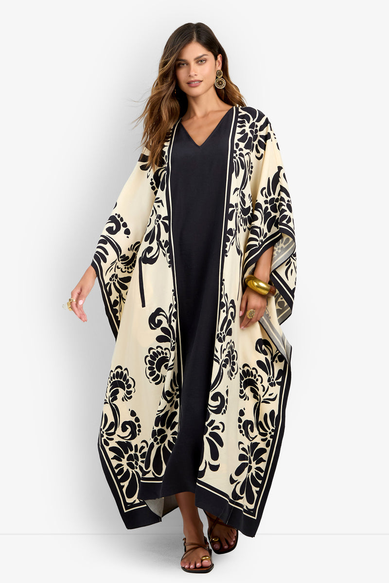 A woman wears the San Martino Caftan—a long black dress with a cream and black floral pattern—styled with gold earrings, bracelets, sandals, and her long hair loose. She stands against a plain white background.