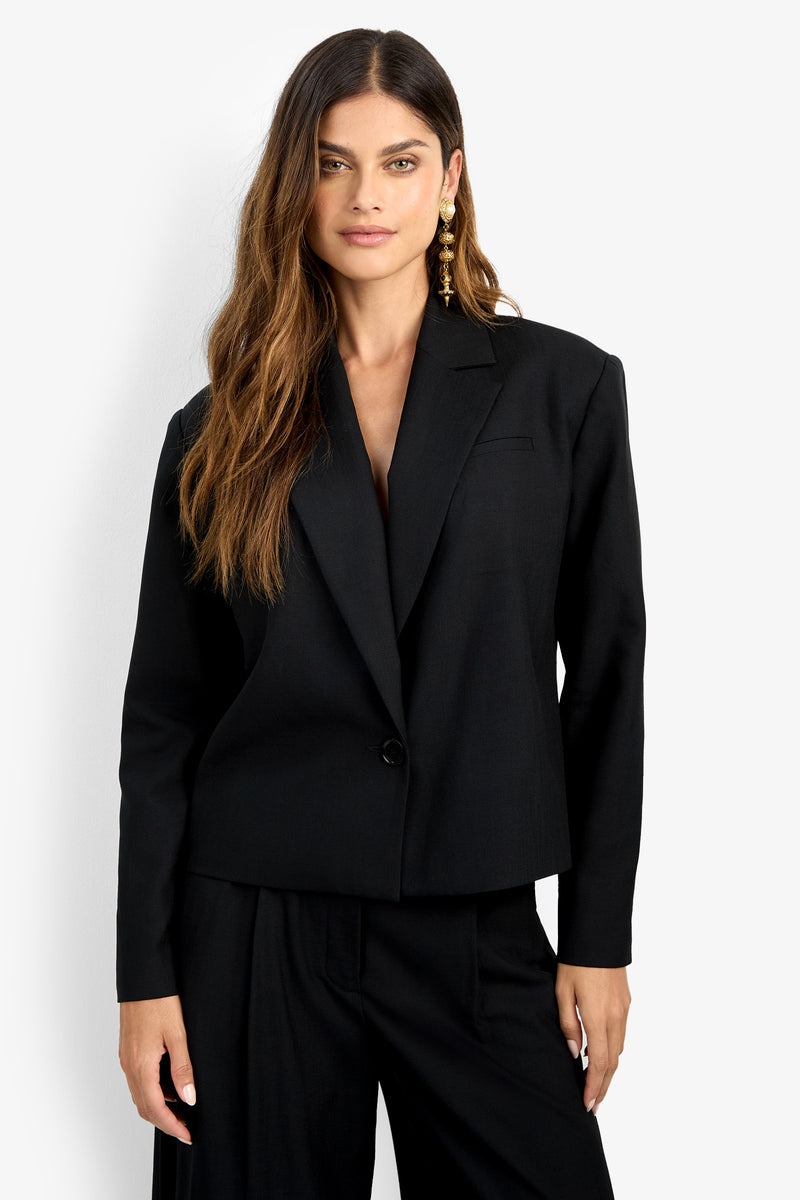 A woman with long wavy hair and gold earrings wears the MALINE JACKET with matching black pants, standing against a white background, facing forward and smiling slightly.