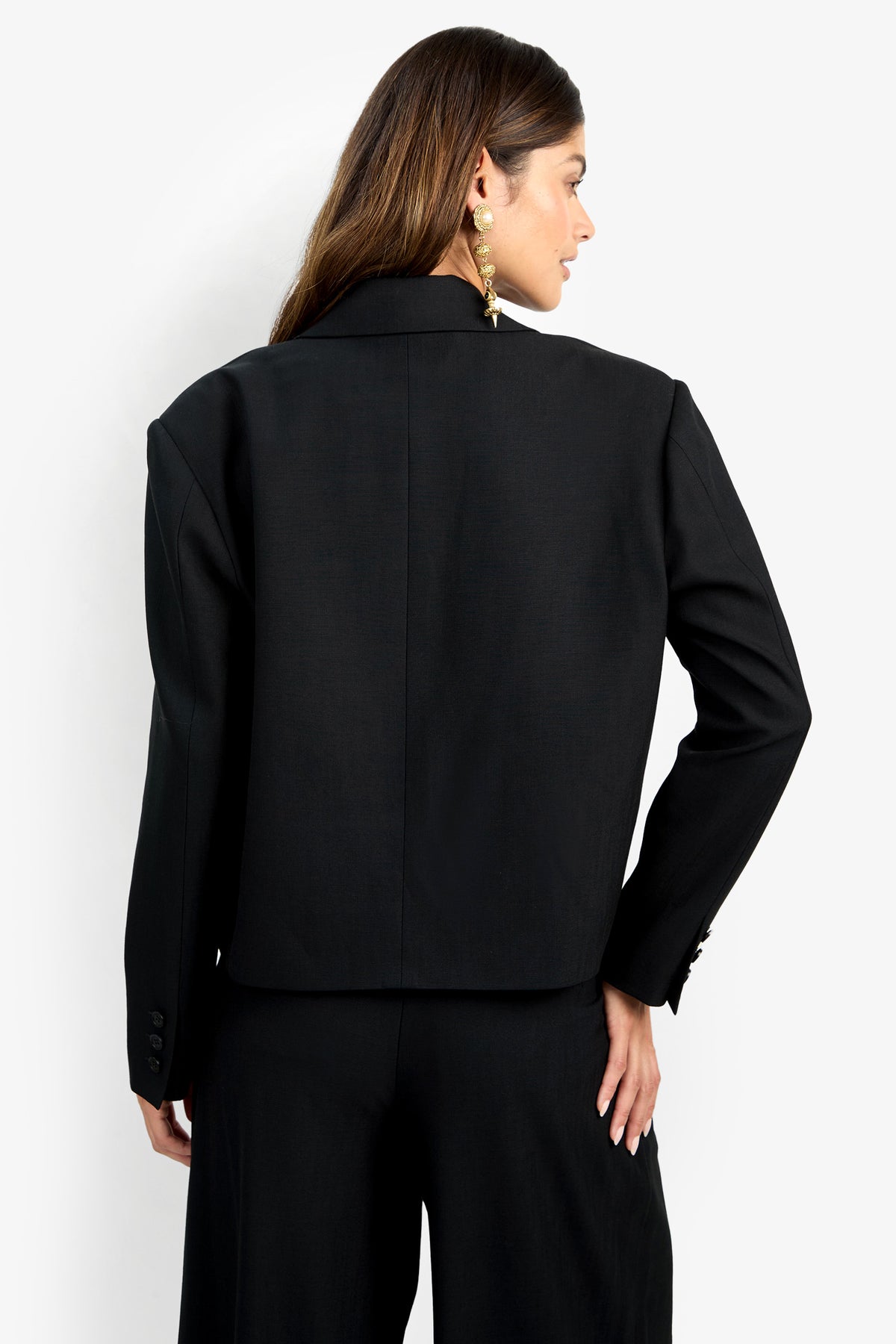 A woman with long brown hair is seen from behind, wearing the MALINE JACKET—a black tailored blazer—with matching pants and large gold earrings. She poses with one hand on her hip against a plain white background.