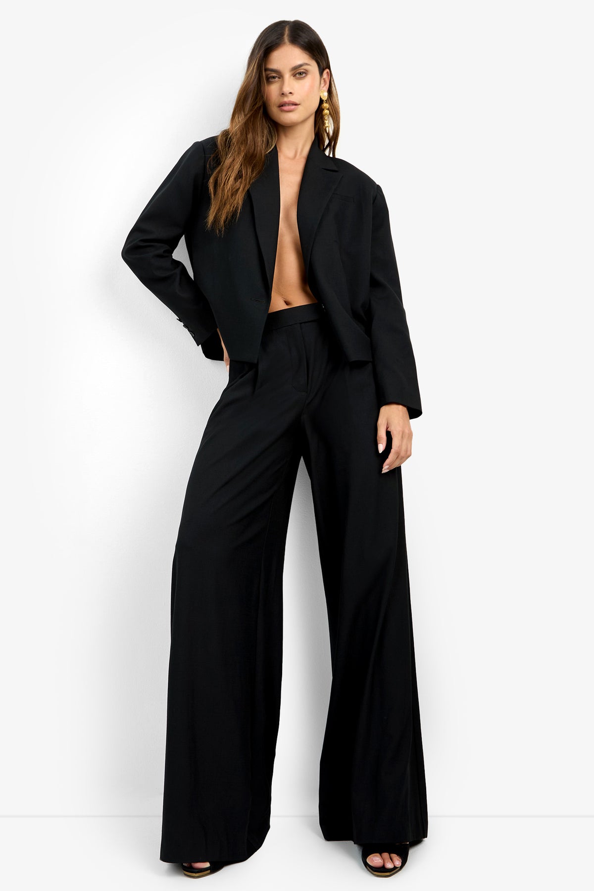 A woman poses confidently against a white background wearing the MIMMO PANT, a loose-fitting black wide-leg trouser. She pairs it with black open-toe heels, gold earrings, and leaves her torso bare beneath the suit jacket.
