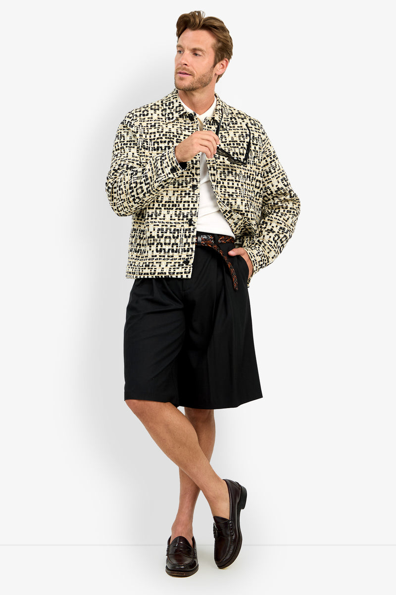 A man with light brown hair poses stylishly against a white background, wearing the PIERRE JACKET over a white shirt, paired with black belted knee-length shorts and dark loafers.