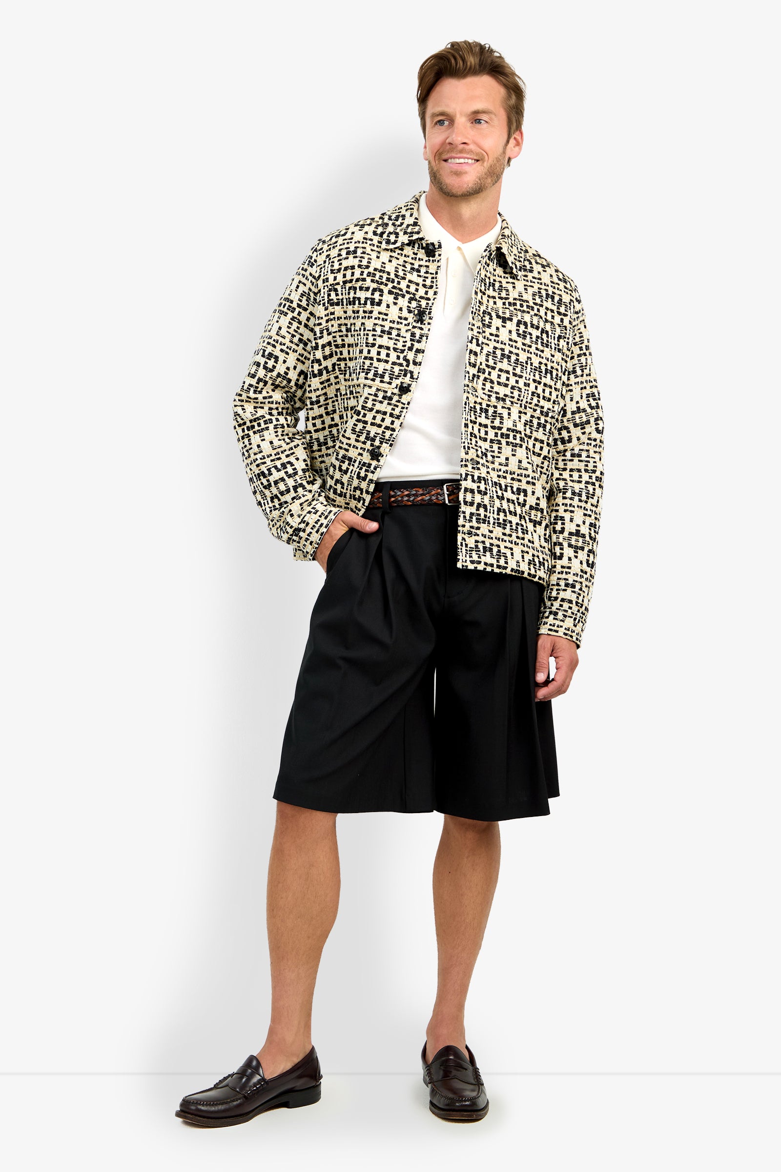 A man smiles against a plain light background, wearing the PIERRE JACKET over a white shirt, paired with black knee-length shorts, a braided belt, and dark loafers.