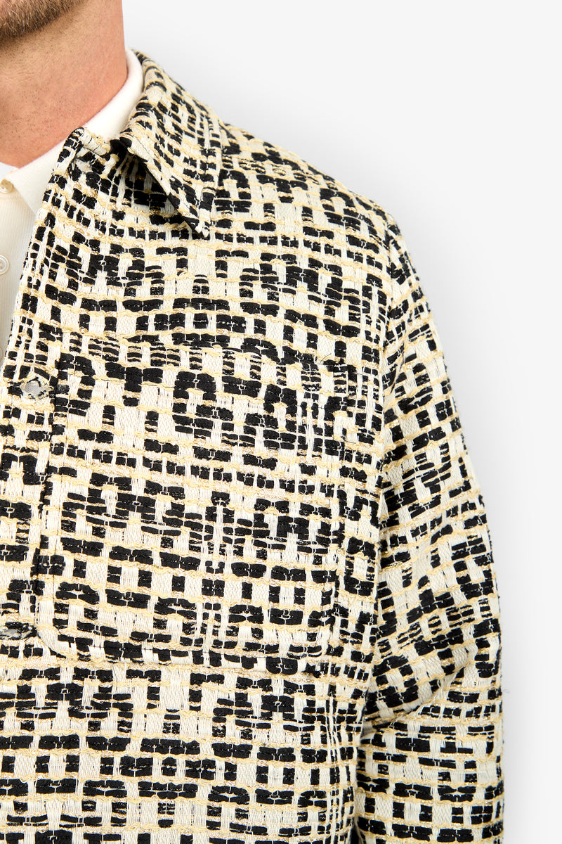 A close-up of someone wearing the PIERRE JACKET with a black and beige geometric texture, a large chest pocket, and a collared neckline. Only part of their face is visible.