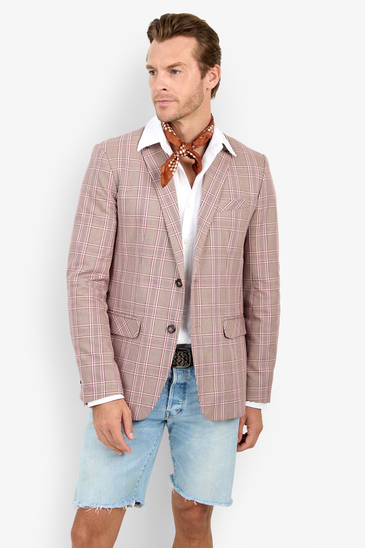 Man wearing the THURSTON BLAZER, white shirt, brown polka dot neck scarf, and light denim shorts stands against a plain background, looking to the side with a neutral expression.