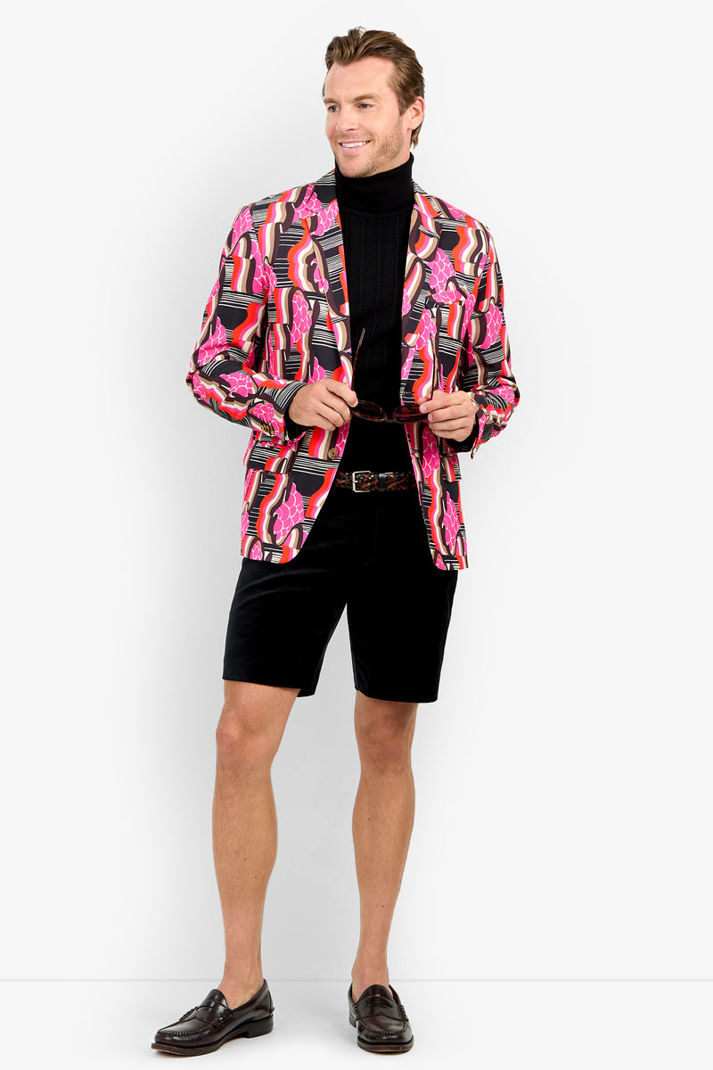 A man smiles as he stands holding open the THURSTON BLAZER, paired with a black turtleneck, black shorts, and brown loafers.