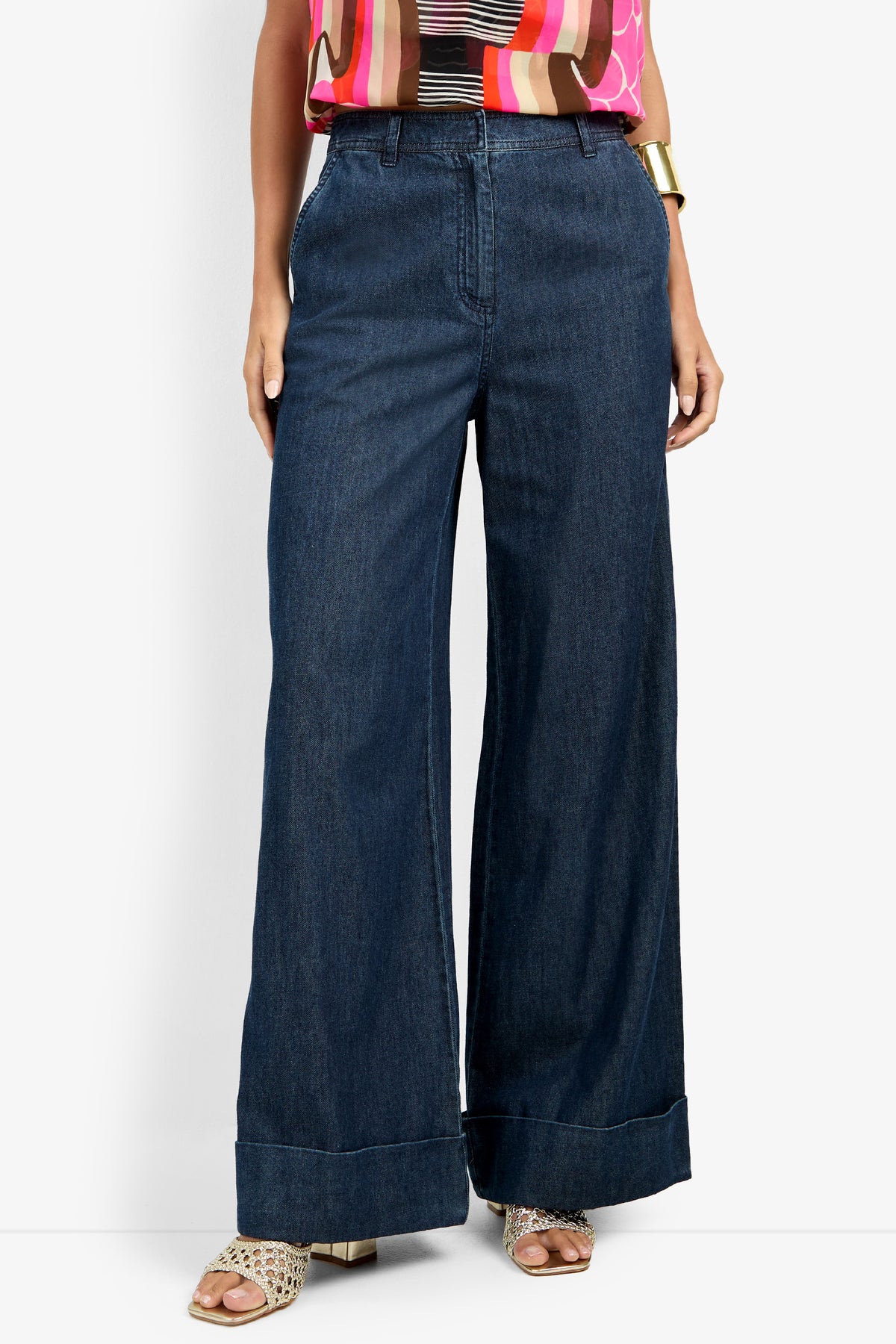 A person wearing DILLAN PANT wide-leg dark blue jeans, a colorful patterned top, a gold bracelet, and open-toe woven sandals stands against a white background.