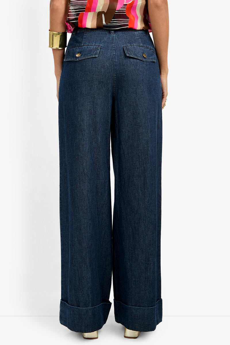 Woman models the DILLAN PANT, featuring high-waisted, wide-leg dark blue jeans with cuffed hems. Seen from the back, she pairs them with a colorful striped top and gold jewelry like a chunky bracelet and ring.