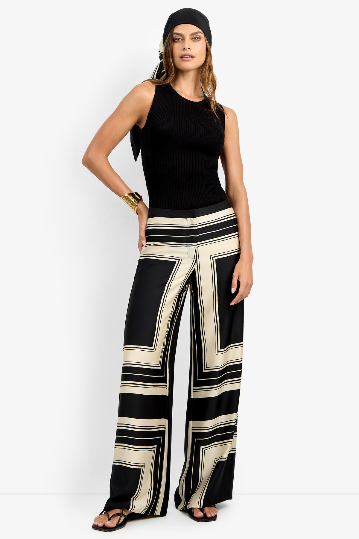 A woman poses confidently against a white background wearing the LANAY PANT, a black sleeveless top, black sandals, a black headscarf, and a gold bracelet.