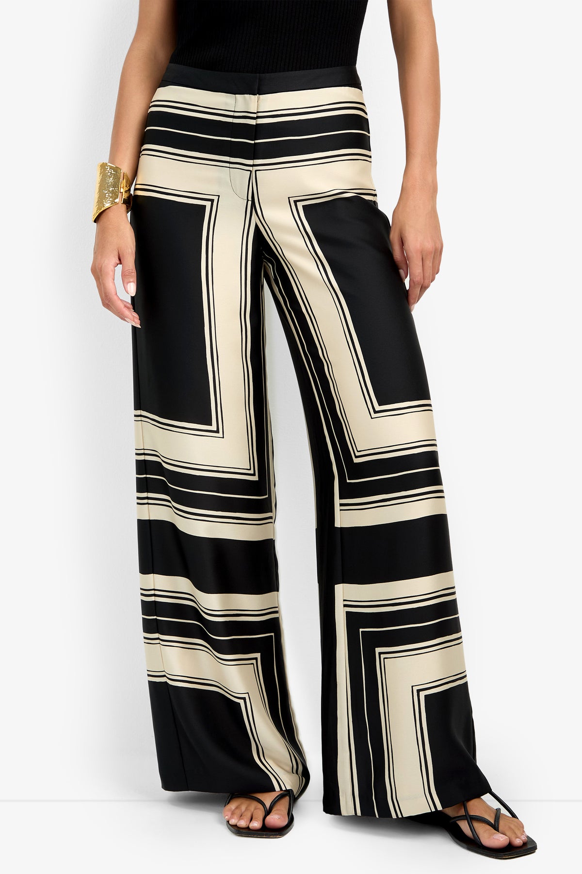 A person wearing the LANAY PANT, featuring a bold black and beige geometric pattern with a wide-leg, high-waisted fit, paired with a black sleeveless top, black sandals, and a large gold cuff bracelet.