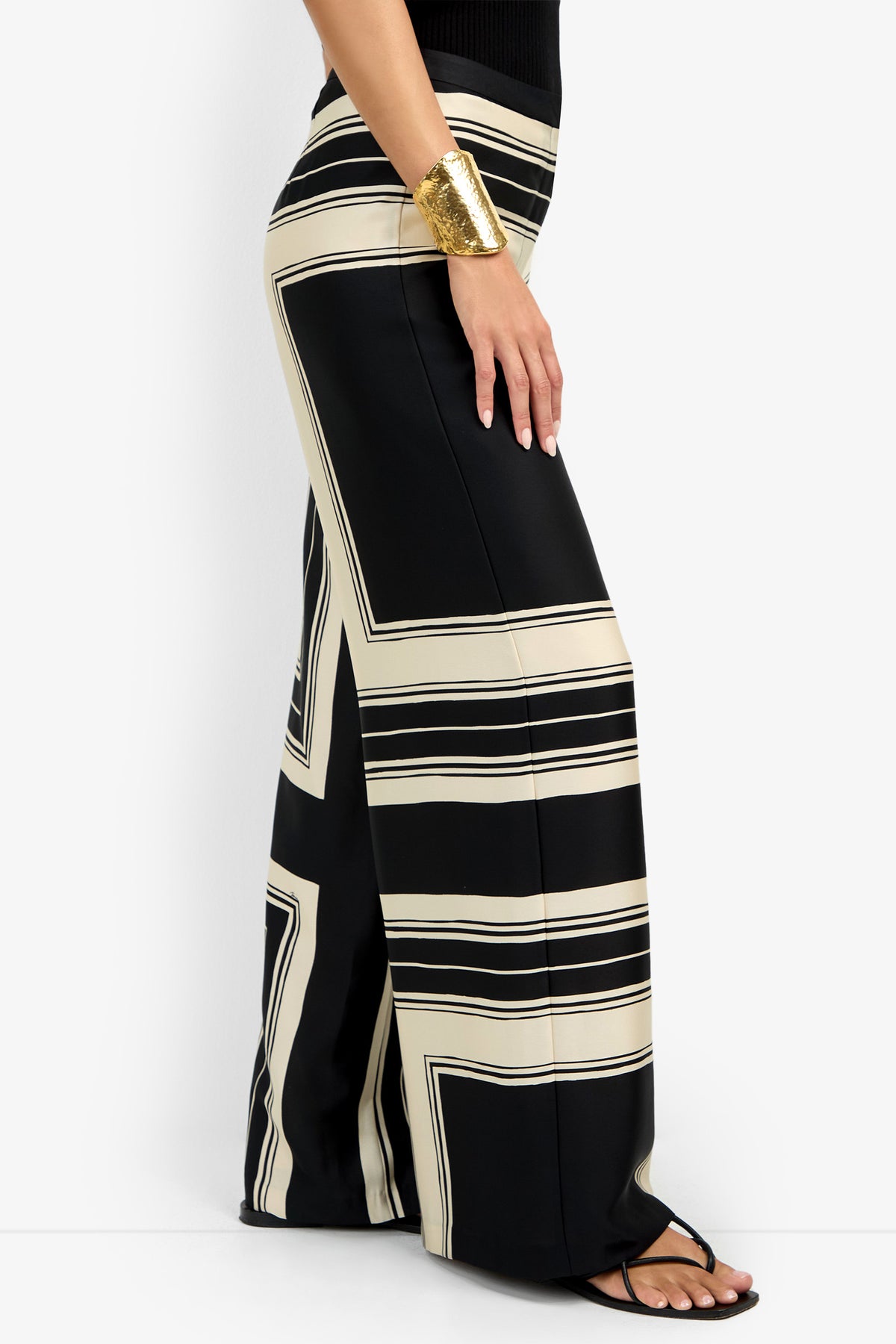 A woman wears the LANAY PANT, featuring high-waisted, wide-leg black pants with bold beige geometric stripes, styled with a sleeveless black top, black sandals, and a large gold cuff bracelet.