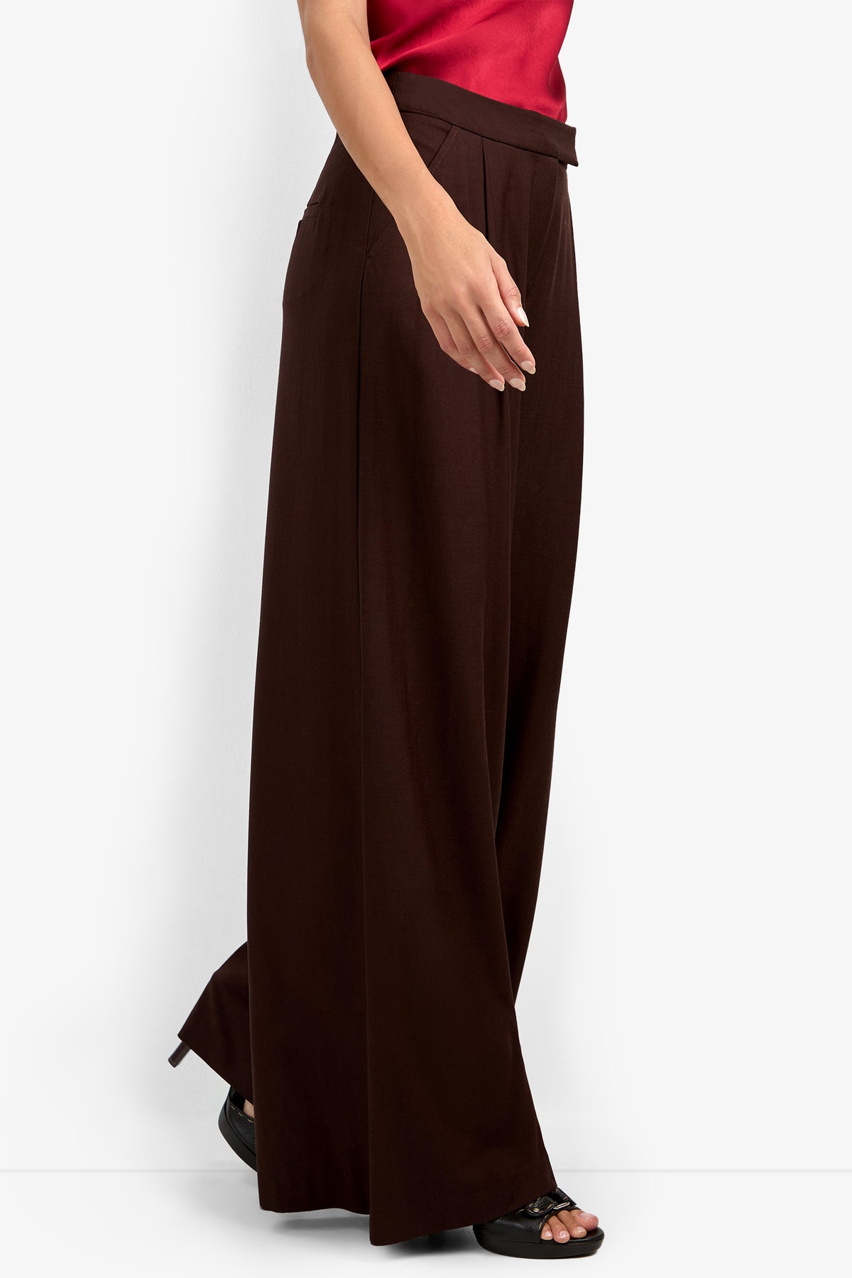 Wearing the MIMMO PANT—wide-leg, high-waisted dark brown pants—with a sleeveless red top and black open-toe sandals, someone walks against a plain white background.