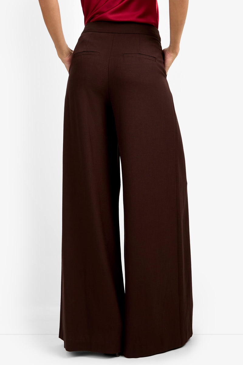 A person is seen from behind, standing with hands on hips, wearing the MIMMO PANT in wide-leg dark brown and a red top, set against a plain white background.
