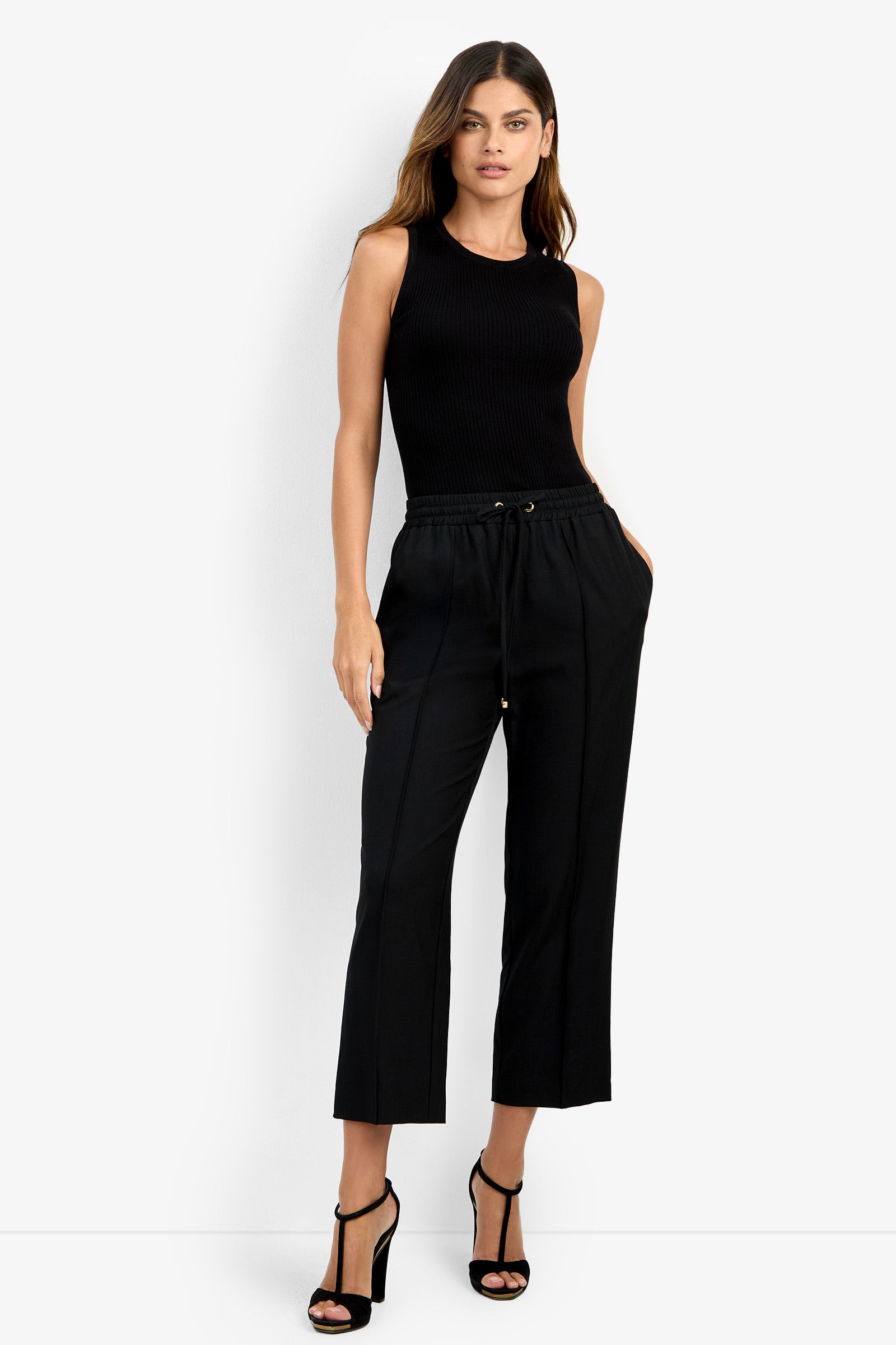 A woman with long, wavy hair stands against a plain background wearing the AVARI PANT, a black drawstring style, paired with a sleeveless black top and black high-heeled sandals, her hands tucked into her pockets.