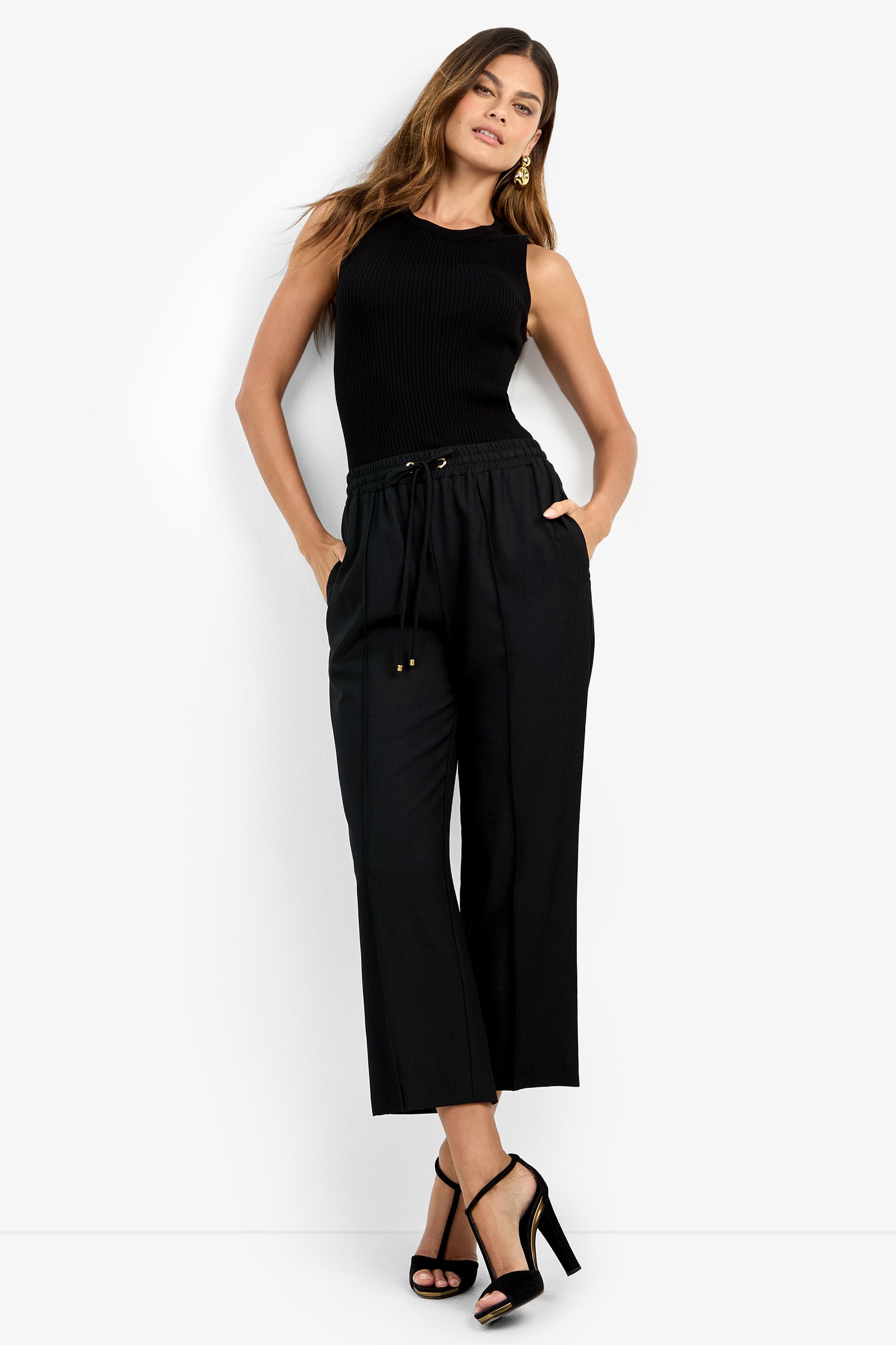 Woman stands against a white background wearing AVARI PANT, a sleeveless black top, and black high-heeled sandals. She has long hair, gold earrings, and her hands are in her pockets.