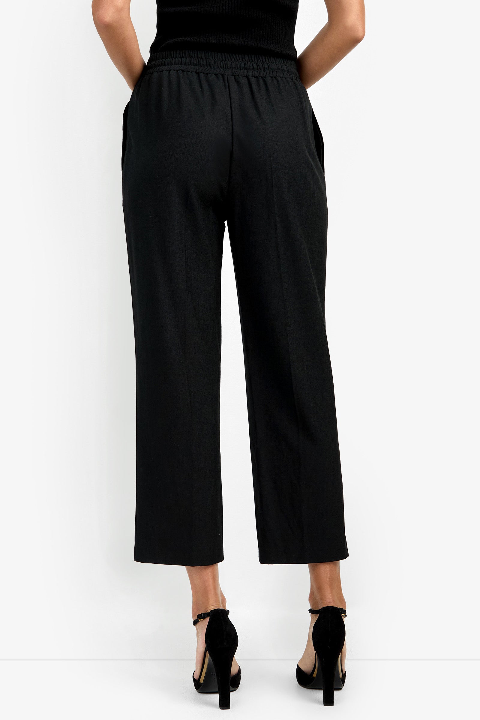 A person wearing the AVARI PANT, a sleeveless black top, and black high-heeled sandals, shown from the back against a plain white background.