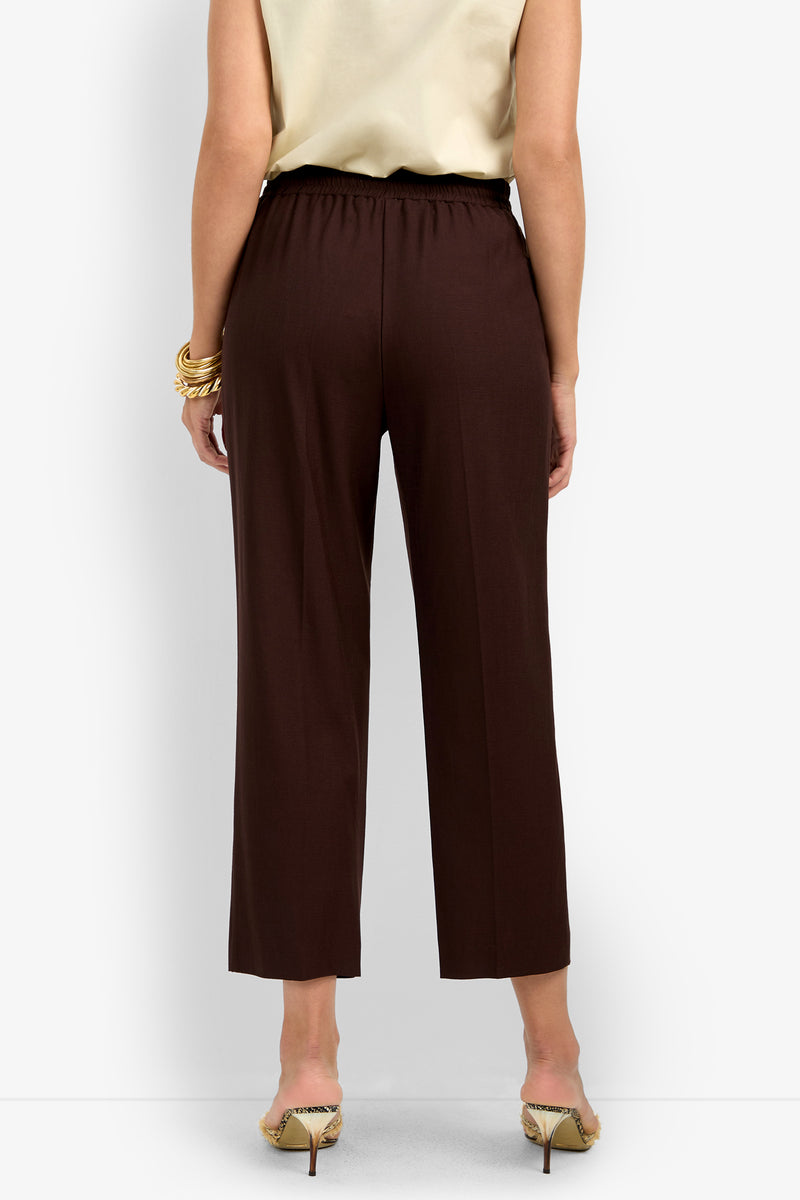 A person stands with their back to the camera wearing a sleeveless beige top, animal print high heels, and AVARI PANT—dark brown wide-leg pants with an elastic waistband.