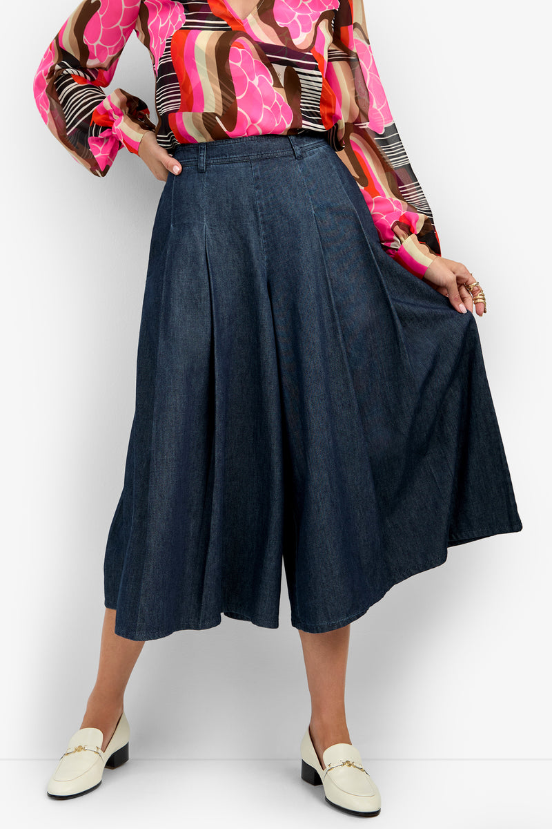 A person wearing the EMELLIO PANT, a pair of blue wide-leg culottes, styled with a pink, brown, and orange patterned blouse and white loafers, stands against a white background while holding out one side of the pants.