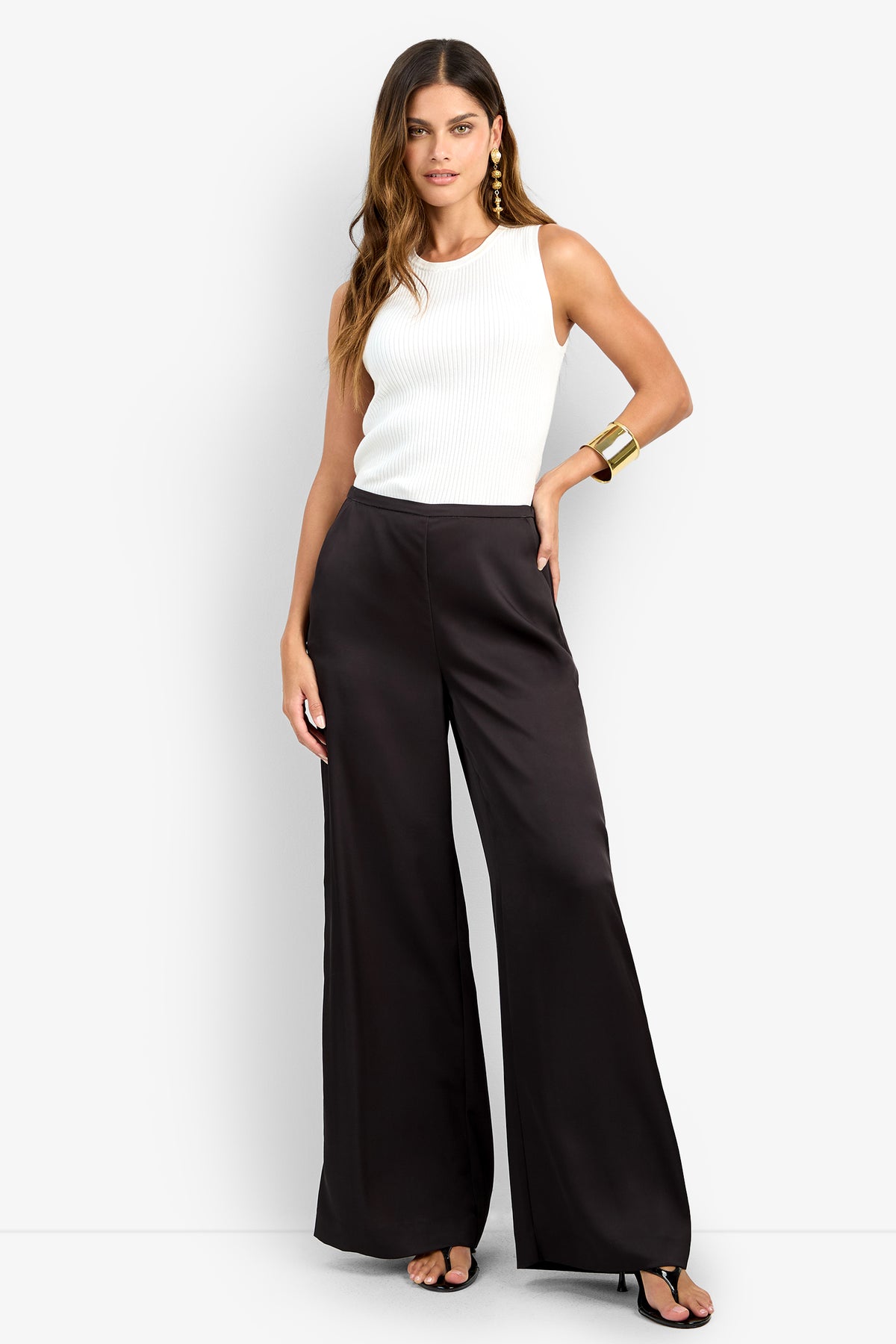 A woman stands against a white background wearing high-waisted black wide-leg TAVIA PANT, a sleeveless white top, black heeled sandals, gold hoop earrings, and a chunky gold bracelet.