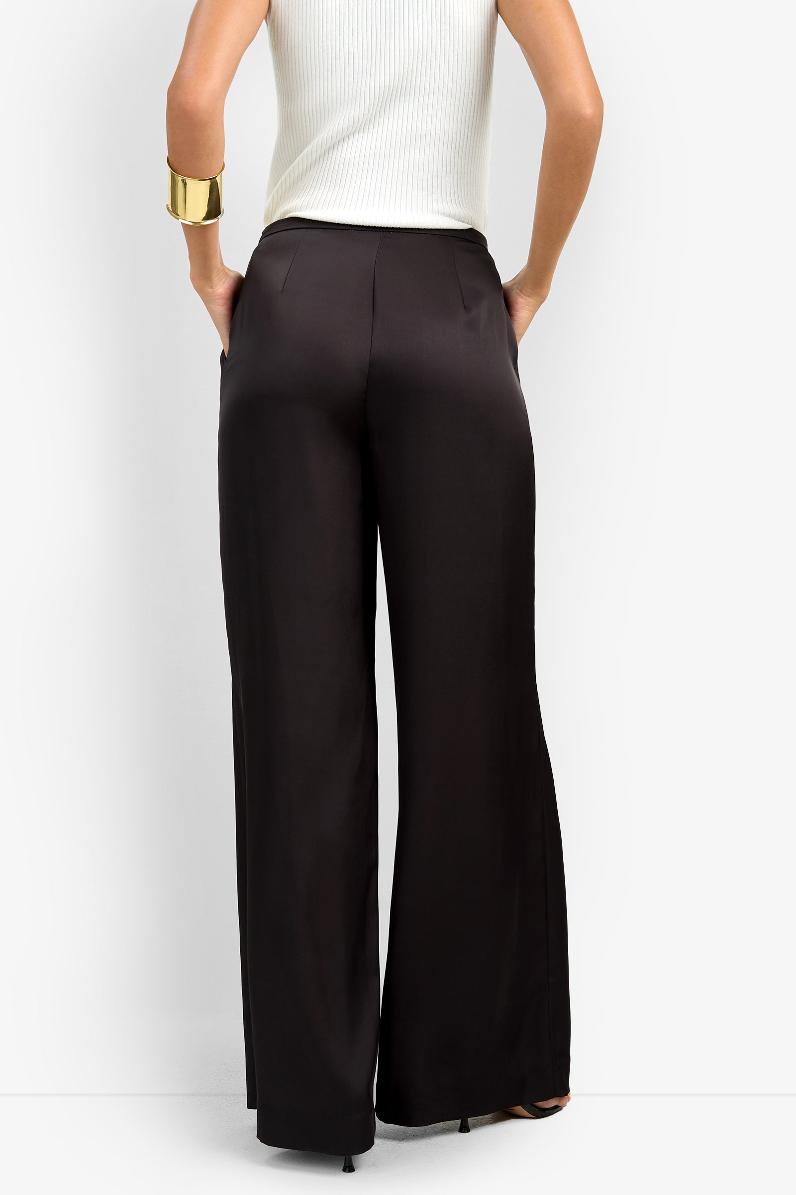 Wearing the high-waisted, wide-leg black TAVIA PANT with a sleeveless white ribbed top, hands in pockets, and a large gold cuff bracelet on the left wrist.