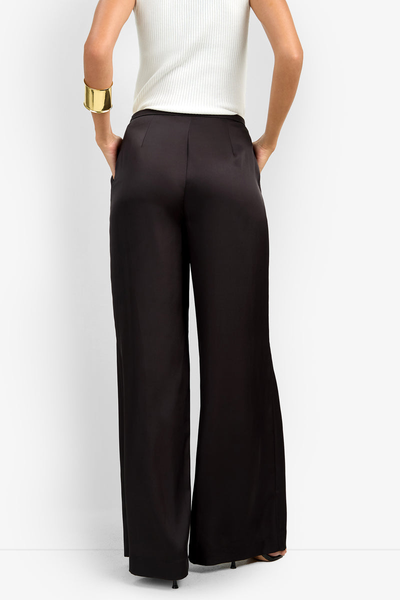 Wearing the high-waisted, wide-leg black TAVIA PANT with a sleeveless white ribbed top, hands in pockets, and a large gold cuff bracelet on the left wrist.