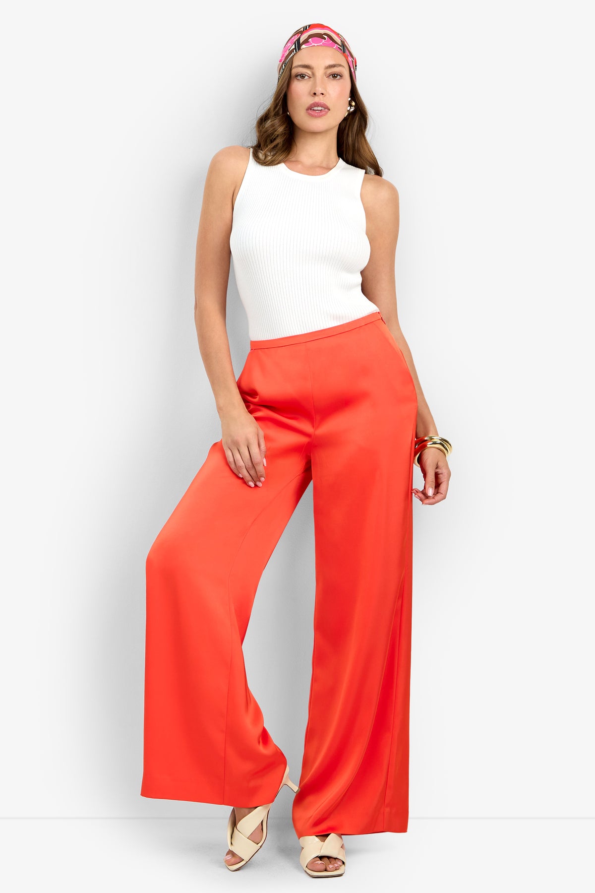 A woman wearing the Tavia Pant in bright orange, a white sleeveless top, beige platform sandals, a pink patterned headband, and a gold bracelet stands against a plain light background.